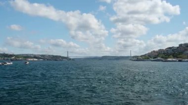 05.14.2025, İstanbul, Türkiye. İstanbul, Türkiye 'deki Boğaz Köprüsü' nün altından geçen büyük gemilerle İstanbul 'da balıkçı tekneleri, manzaralı bulutlar, tepeler ve arka planda bir Türk bayrağıyla aydınlık bir günde