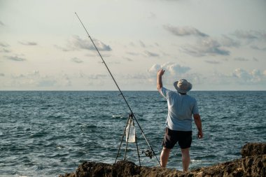 Kayalık kıyı şeridinde oturmuş oltayla balık tutarken açık güneşli gökyüzü altında sakin deniz dalgalarını seyreden adam Akdeniz kıyısı açıkhava aktivitesini rahatlatıyor.