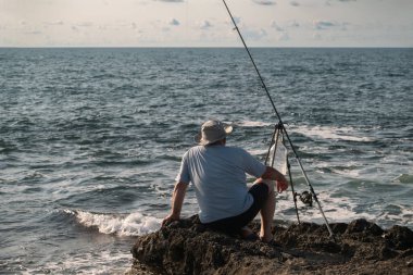 Kayalık kıyı şeridinde oturmuş oltayla balık tutarken açık güneşli gökyüzü altında sakin deniz dalgalarını seyreden adam Akdeniz kıyısı açıkhava aktivitesini rahatlatıyor.