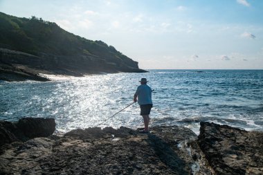 Kayalık kıyı şeridinde oturmuş oltayla balık tutarken açık güneşli gökyüzü altında sakin deniz dalgalarını seyreden adam Akdeniz kıyısı açıkhava aktivitesini rahatlatıyor.