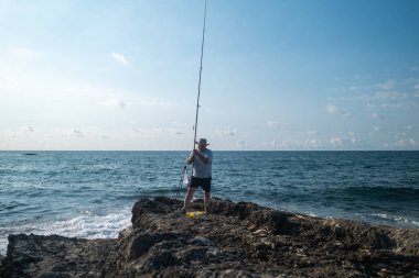 Güneşli bir günde açık mavi gökyüzü ve okyanus dalgalarıyla kayalık sahil şeridinde balıkçılık - açık hava eğlencesi ve deniz aktivitesi