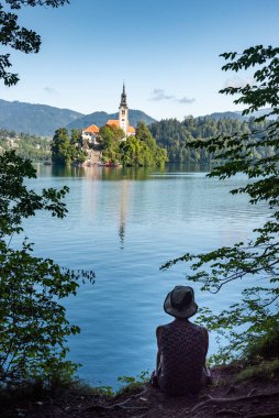 Landscape of Slovenia. The silhouette of a seated person stands out from the blue waters of Lake Bled. In the background, we can see The Church of the Mother of God, also called the Pilgrimage Church of the Assumption of Mary clipart