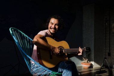 Young Hispanic man playing acoustic guitar sitting on floor in living room at Sunny Stylish Loft Apartment in Mexico Latin America