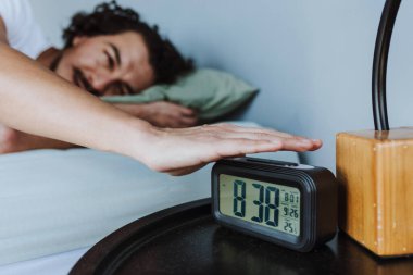 Hispanic young man in bed reaching for alarm clock on bedside table at home in Mexico Latin America
