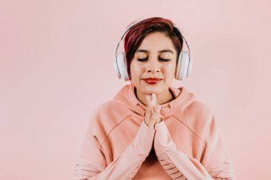 young hispanic woman girl listening music with headphones  and holding mobile phone on coral pink background with copy space studio portrait in Mexico Latin America