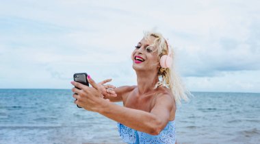 Makyaj yapan genç transseksüel adam ve kadın kıyafetleri Meksika 'da Latin Amerika plajında kulaklık ve cep telefonuyla müzik dinliyor.