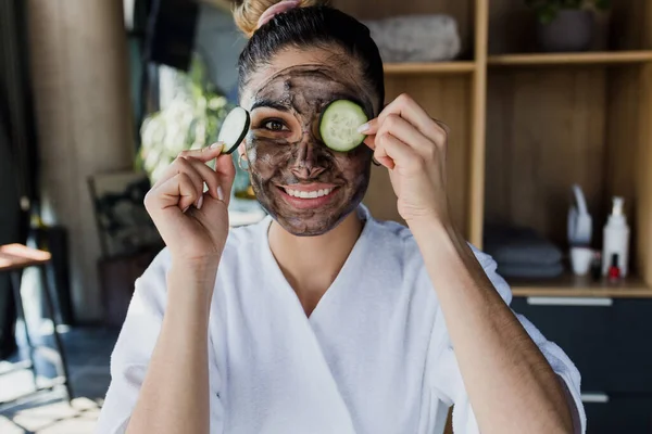 young latin woman with facial mask on her face for Skin care holding slices of cucumber at home in Mexico, Hispanic female