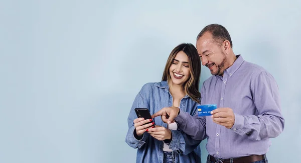 latin father and daughter in casual clothes holding mobile phone and credit card on blue background in Mexico Latin America