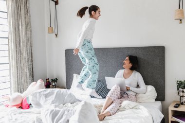 Latin anne yatakta yatıp dizüstü bilgisayarıyla çalışıyor ve kızı Meksika Latin Amerika 'da ofis kavramında yatakta zıplıyor. İspanyol kadın aile.
