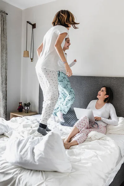 latin mother lying on bed and working with her laptop and daughter jumping on bed at home office concept in Mexico Latin America, hispanic female family