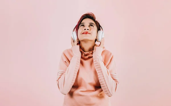 young latin woman girl listening music with headphones  and holding mobile phone on coral pink background with copy space studio portrait in Mexico Latin America, hispanic people