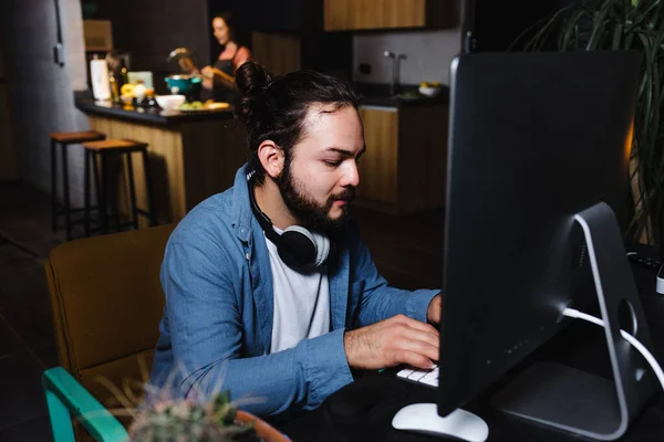 Young latin man working and using computer while girlfriend is cooking on background at home at night in Mexico Latin America, hispanic people