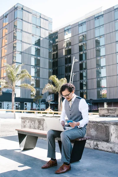 young latin businessman using laptop and phone working on computer in bench outdoors in Mexico city, Latin America hispanic people