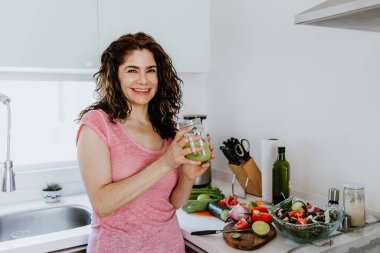 Latin kadın yeşil meyve suyu içiyor ve Meksika 'da evdeki mutfakta malzemeleri işliyor.