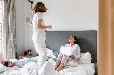 Latin anne yatakta yatıp dizüstü bilgisayarıyla çalışıyor ve kızı Meksika Latin Amerika 'da ofis kavramında yatakta zıplıyor. İspanyol kadın aile.