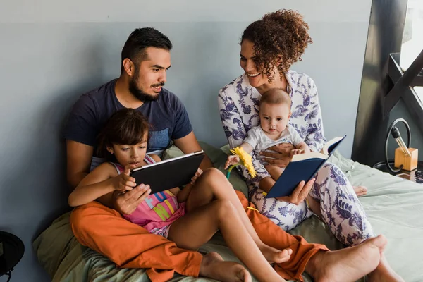 young latin family with two children reading a book together on bed at home in Mexico Latin America, hispanic mother and father