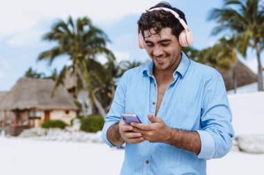 Latin Amerika, Karayipler ve tropikal bölgelerde tatillerde veya tatillerde kulaklık takan ve telefonda müzik dinleyen İspanyol genç adam.