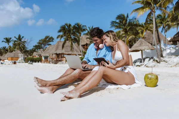 hispanic young couple of digital nomads sit down on the beach with laptop and tablet enjoying the travel adventure vacation and working in Mexico Latin America, Caribbean and Tropical destinations