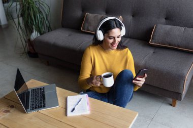 Latin yetişkin bir kadın Meksika 'da evinde telefon ve kulaklık kullanırken kanepede oturuyor. Orta yaşlı Latin bir kadın.