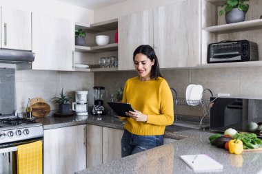 Latin yetişkin kadın sağlıklı yemekler pişiriyor ve Meksika Latin Amerika 'da evdeki mutfakta dijital tablet tutuyor.