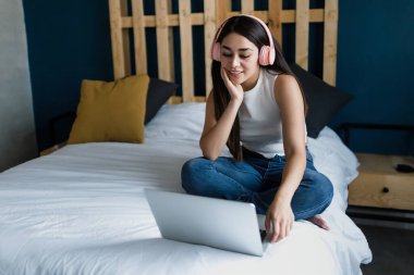 Latin Amerika 'da Latin kökenli Latin kökenli genç bir kız öğrenci, evinde yatağında dizüstü bilgisayar ve kulaklık kullanıyor.