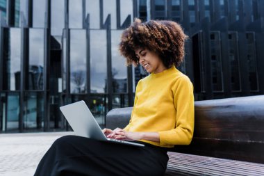 Latin Amerika şehrinde dizüstü bilgisayar kullanan genç Afrikalı Amerikalı iş kadını, gökdelen geçmişe sahip İspanyol finans ve Karayipliler