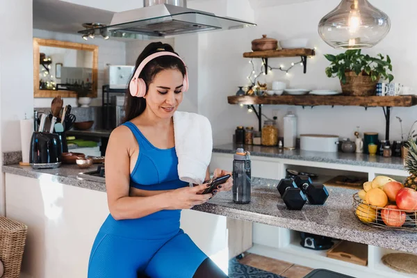 young latin woman resting after workout at home using mobile phone and headphones at kitchen in Home in Mexico Latin America, hispanic female in wellness concept