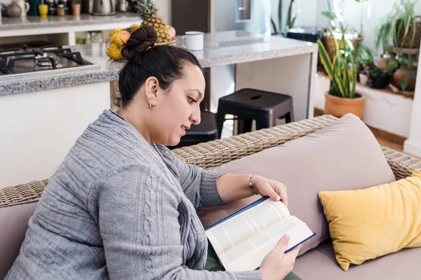 Young latin woman overweight reading book on sofa at home in Mexico Latin America, hispanic female plus size student