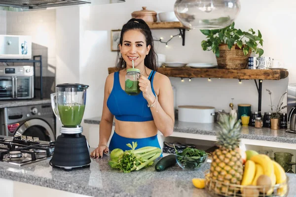 Latin woman pouring green juice from mixer into glass in kitchen at home in Mexico Latin America, hispanic female on detox diet