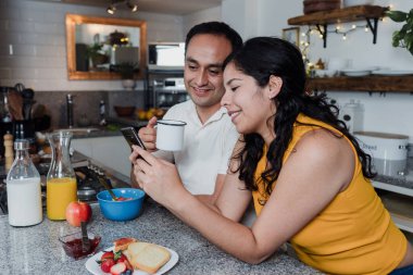 Latin çift kahvaltı yapıyor ve Meksika 'da mutfakta cep telefonu kullanıyor. Latin Amerikalılar eğleniyor.