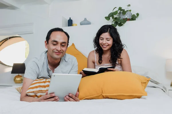 Latin couple reading book and using tablet in bed at home in Mexico Latin America, hispanic people having fun