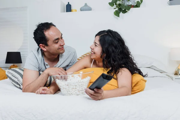 Latin couple watching a movie lying on bed at home in Mexico Latin America, hispanic people eating popcorn on bedroom