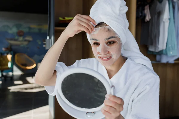 young latin woman with facial mask on her face for Skin care looking a mirror at home in Mexico Latin America, hispanic female health care