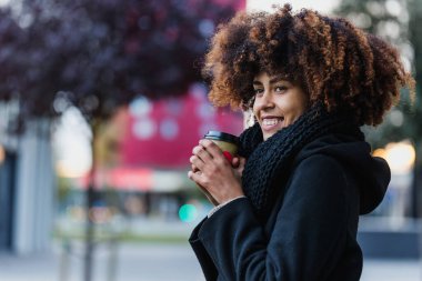 Latin Amerika şehrinde kahve fincanı tutan genç Afrikalı Amerikalı iş kadını, gökdelen geçmişe sahip İspanyol finans ve Karayipliler.