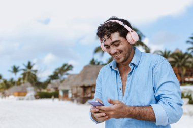 Latin Amerika, Karayipler ve tropikal bölgelerde tatillerde veya tatillerde kulaklık takan ve telefonda müzik dinleyen İspanyol genç adam.