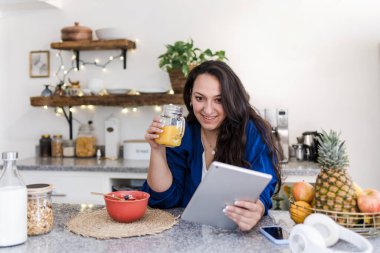 Latin Amerika 'da ev mutfağında kahvaltı ederken tablet kullanan aşırı kilolu Latin bir kadın. Latin kökenli, iri yarı bir kadın.