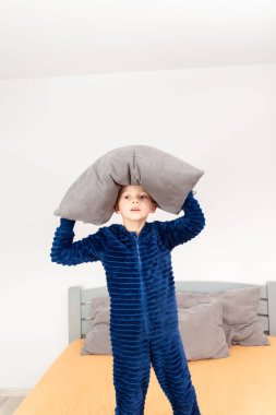 A six-year-old boy of European appearance is dressed in blue pajamas. The boy is waving and hugging with a gray pillow.