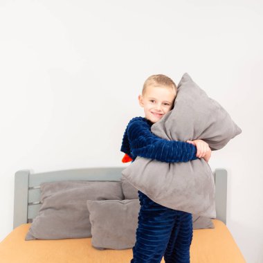 A six-year-old boy of European appearance is dressed in blue pajamas. The boy is waving and hugging with a gray pillow.