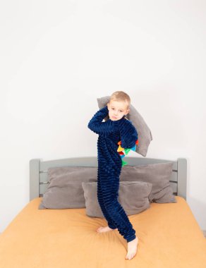 A six-year-old boy of European appearance is dressed in blue pajamas. The boy is waving and hugging with a gray pillow.