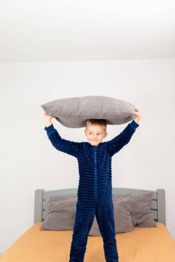 A six-year-old boy of European appearance is dressed in blue pajamas. The boy is waving and hugging with a gray pillow.