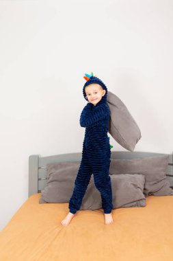 A six-year-old boy of European appearance is dressed in blue pajamas. The boy is waving and hugging with a gray pillow.