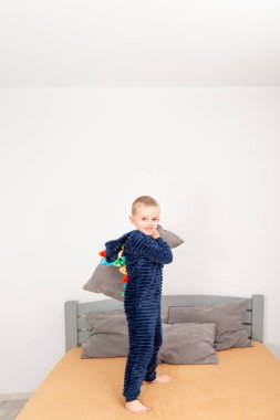 A six-year-old boy of European appearance is dressed in blue pajamas. The boy is waving and hugging with a gray pillow.