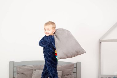 A six-year-old boy of European appearance is dressed in blue pajamas. The boy is waving and hugging with a gray pillow.