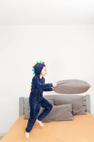 A six-year-old boy of European appearance is dressed in blue pajamas. The boy is waving and hugging with a gray pillow.