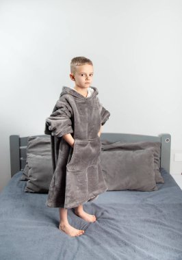 A six-year-old boy of European appearance is dressed in gray pajamas, jumping and having fun on a gray bed.