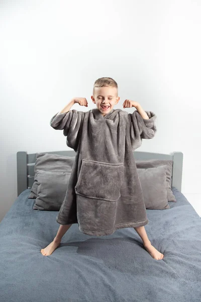 A six-year-old boy of European appearance is dressed in gray pajamas, jumping and having fun on a gray bed.
