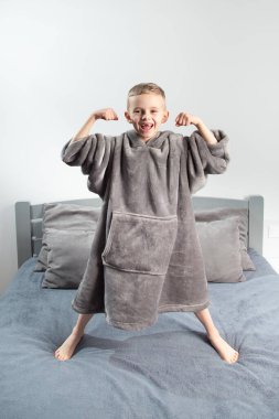 A six-year-old boy of European appearance is dressed in gray pajamas, jumping and having fun on a gray bed.