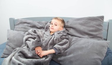 A six-year-old boy of European appearance is dressed in gray pajamas, lying and having fun on a gray bed.