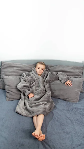 A six-year-old boy of European appearance is dressed in gray pajamas, lying and having fun on a gray bed.