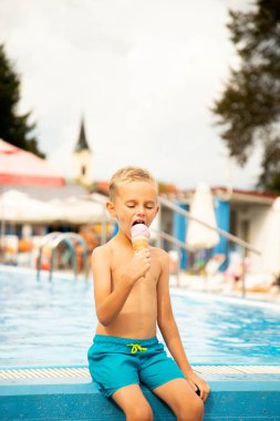 Yedi yaşında Avrupalı görünümlü bir çocuk havuzun kenarında oturur ve dondurma yer..